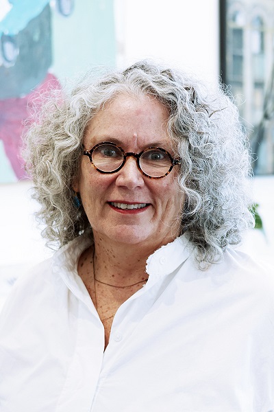 A woman with curly gray hair wearing a white shirt smiles at the camera.